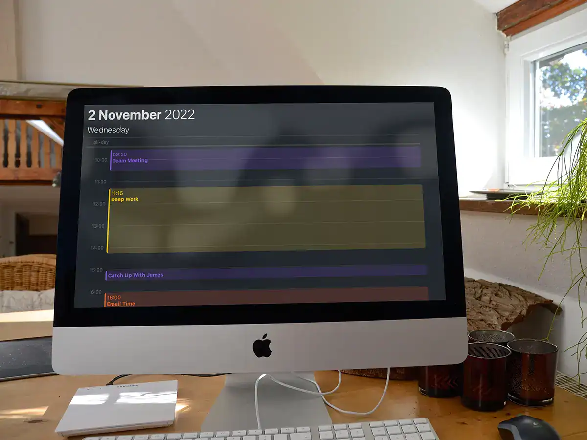 Professional iMac on a wooden desk displaying a digital schedule for November 2, 2022, with appointments like team meetings, deep work, and catch-up calls, situated in a cozy, well-lit room with a window and houseplant.