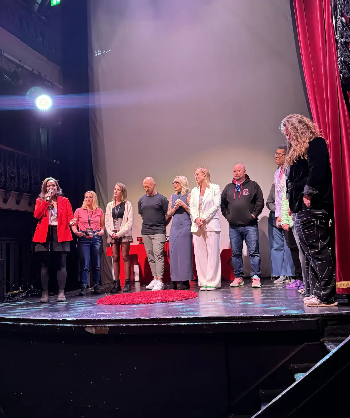 Women speaking at a TedX event on stage with diverse speakers, one woman in a red blazer addressing the audience, TEDx logo on stage, with red curtains and stage lighting.